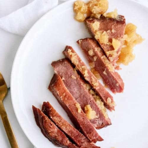 Sliced cooked ham topped with pineapple chunks arranged on a white plate, next to a gold fork and knife on a white surface.