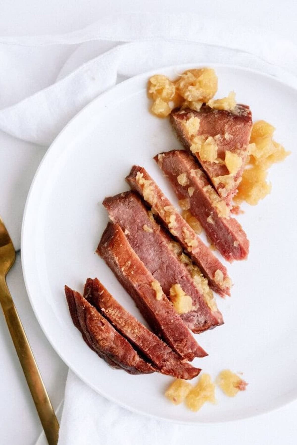 Sliced cooked ham topped with pineapple chunks arranged on a white plate, next to a gold fork and knife on a white surface.