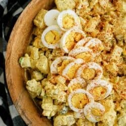 A wooden bowl filled with potato salad topped with sliced boiled eggs and sprinkled with paprika. A black and white checkered cloth is partially visible underneath.