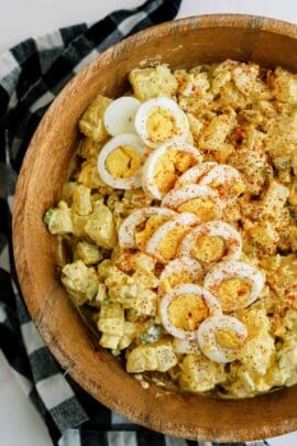 A wooden bowl filled with potato salad topped with sliced boiled eggs and sprinkled with paprika. A black and white checkered cloth is partially visible underneath.