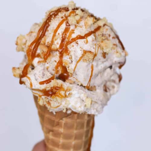 A hand holds a waffle cone filled with ice cream topped with caramel drizzle and crumbly bits against a plain white background.