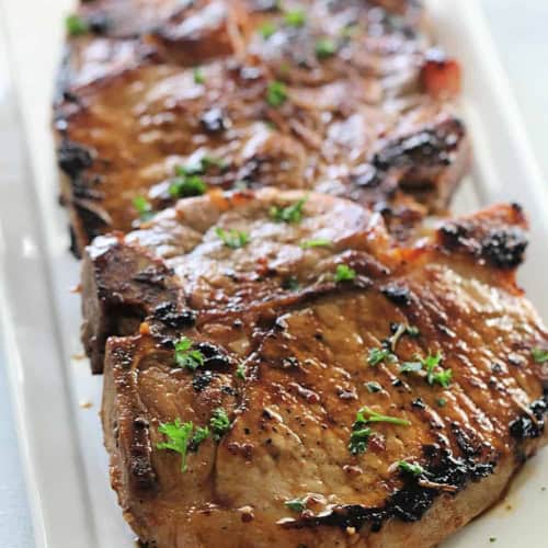 Three grilled pork chops garnished with chopped parsley are arranged on a rectangular white plate.