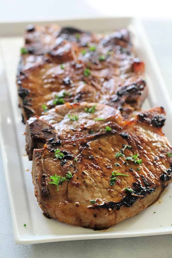 Three grilled pork chops garnished with chopped parsley are arranged on a rectangular white plate.