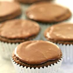 Six chocolate cupcakes in silver foil wrappers, each topped with a smooth layer of chocolate frosting, arranged in a soft-focus background.
