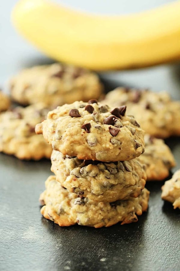 A stack of 3 Chocolate Chip Banana Oatmeal Cookies surrounded by other Chocolate Chip Banana Oatmeal Cookies and a banana in the background.