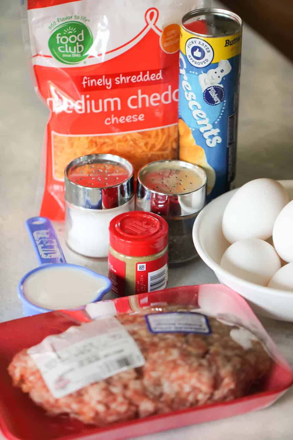 Ingredients for a recipe: shredded cheddar cheese, crescent rolls, ground meat, eggs, seasoning, milk, salt, and pepper on a kitchen counter.