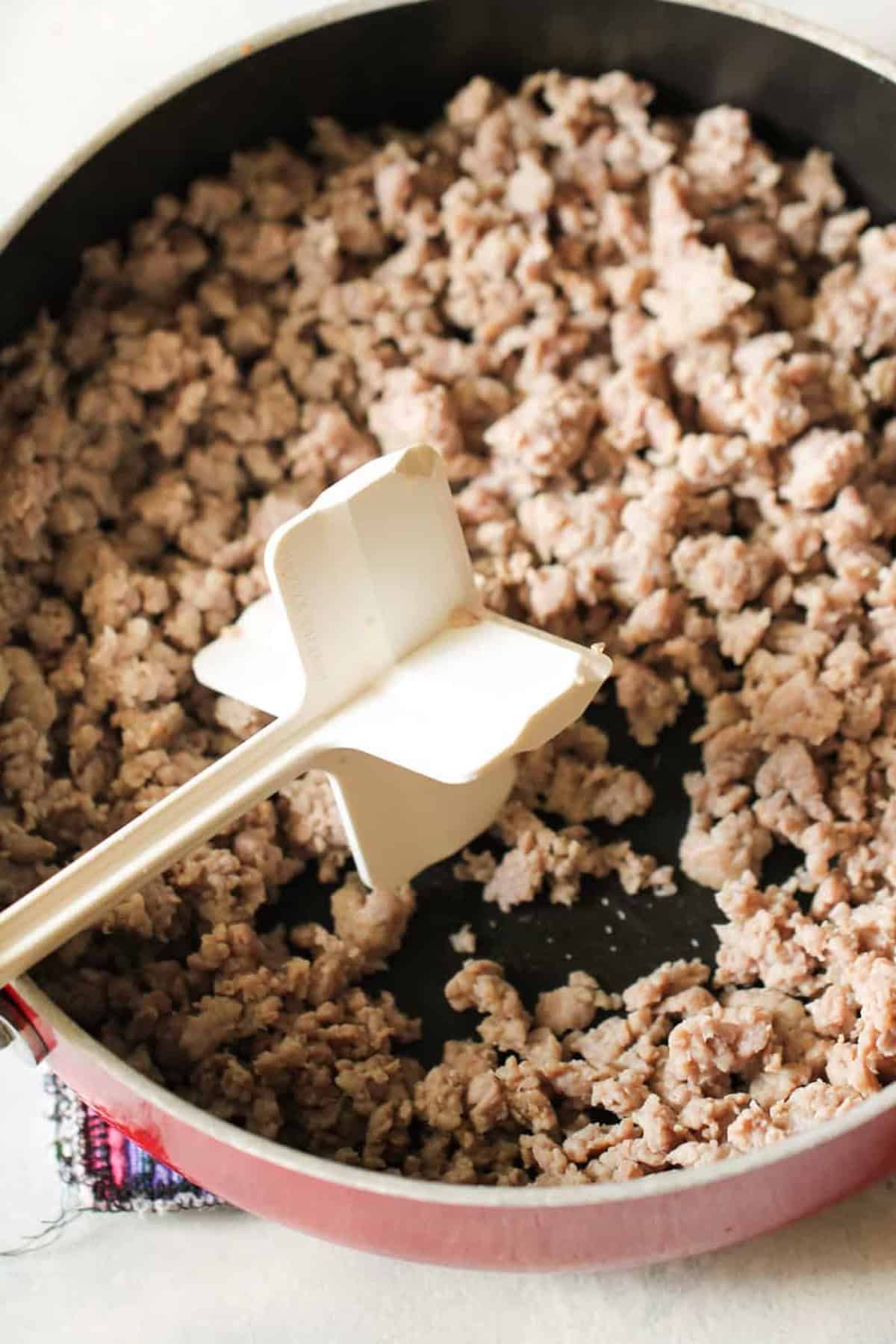 Ground meat is being browned in a frying pan with a meat chopping tool visible.