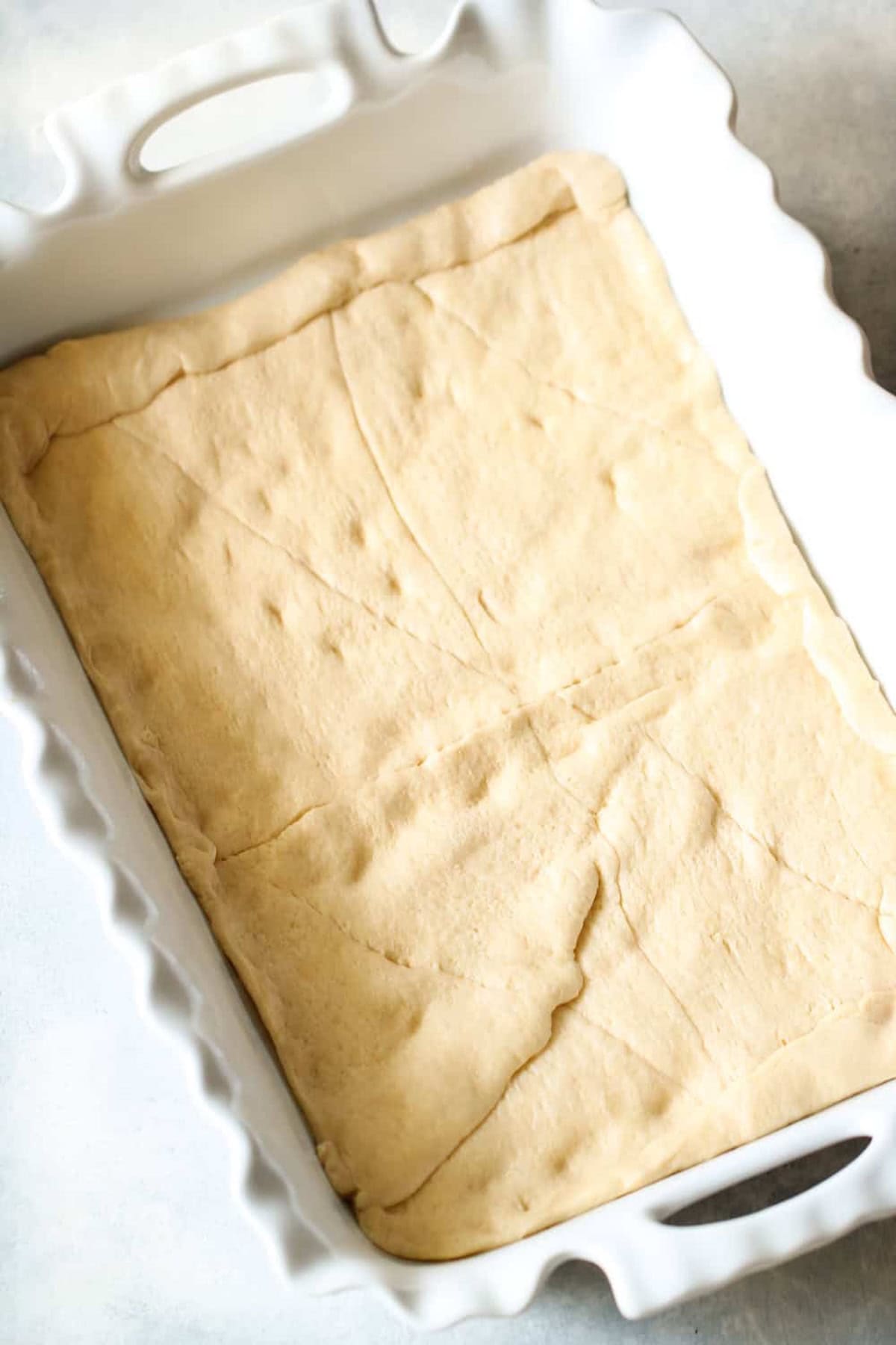 A white rectangular baking dish with uncooked dough pressed evenly at the bottom, showing slight creases and folds.
