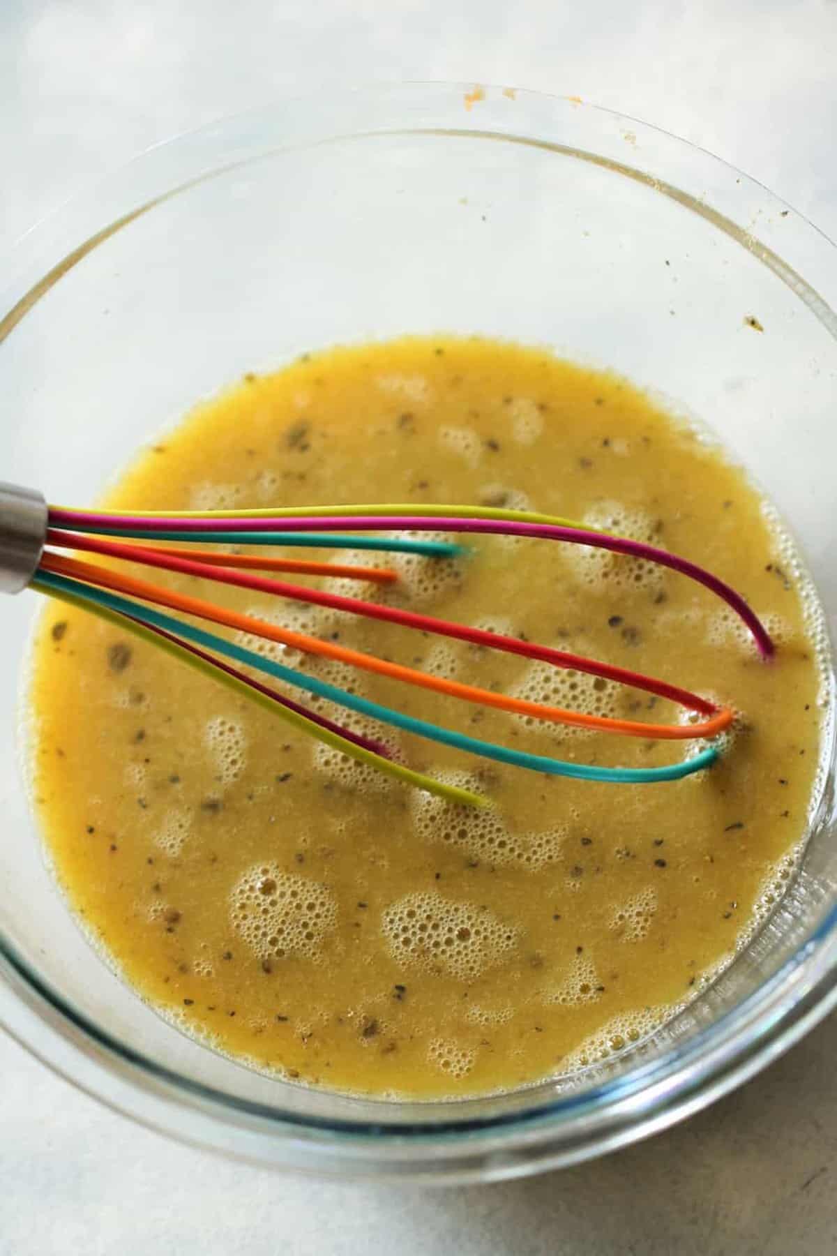 A glass bowl containing a yellow liquid mixture with herbs is being stirred by a colorful whisk.