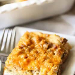 A slice of cheesy breakfast casserole on a white plate with a fork, featuring a browned top and visible bits of sausage and eggs.