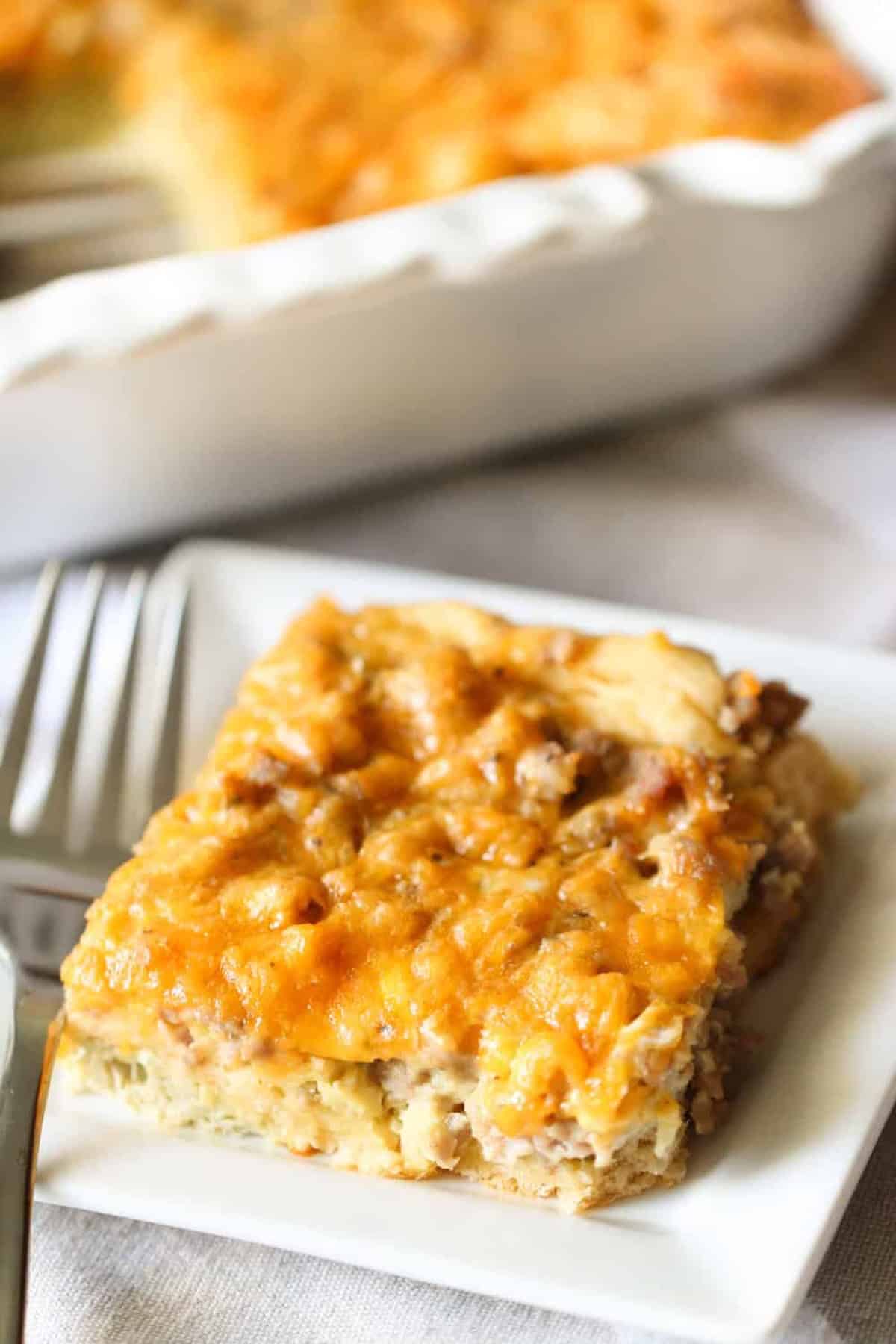 A slice of cheesy breakfast casserole on a white plate with a fork, featuring a browned top and visible bits of sausage and eggs.
