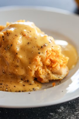 A piece of baked chicken topped with a thick layer of cheese and creamy gravy, garnished with black pepper, served on a white plate.