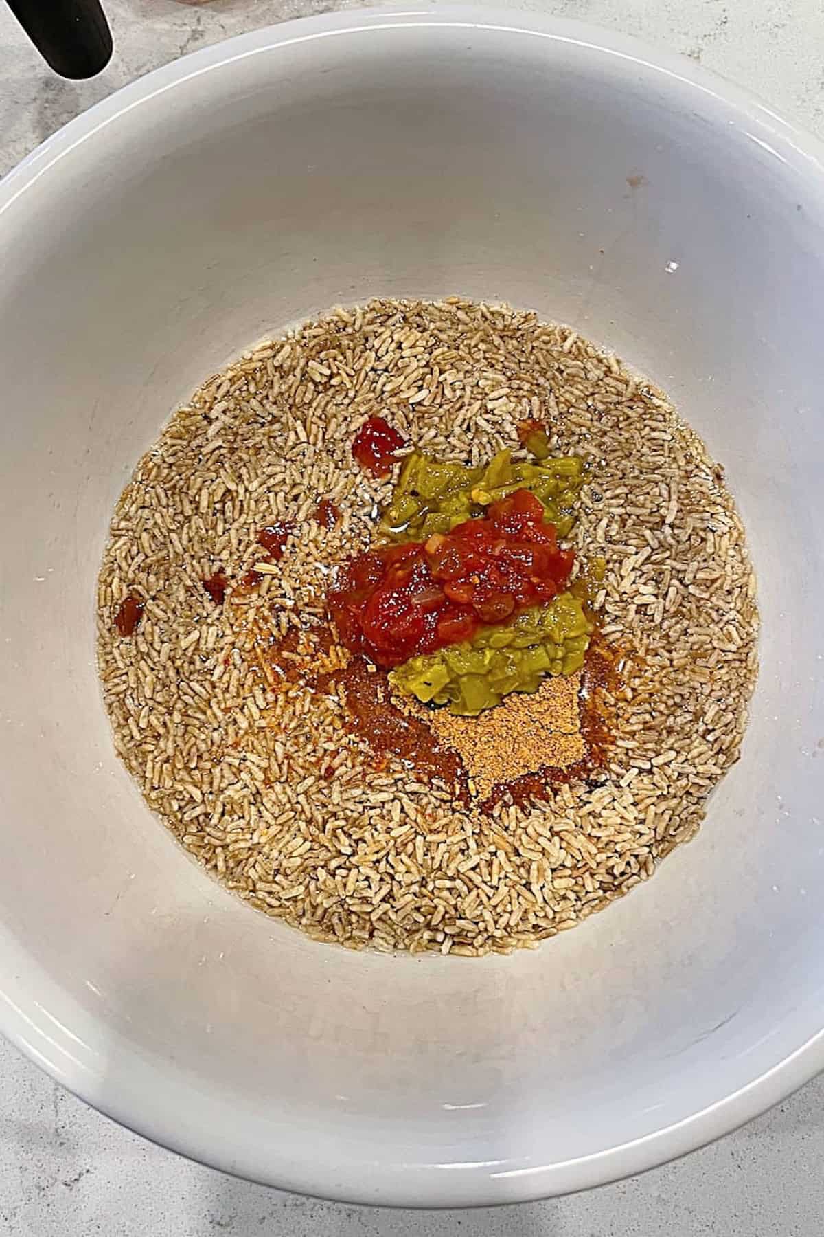 A white bowl contains uncooked brown rice, canned diced tomatoes, chopped green chiles, and various seasonings on a light countertop.