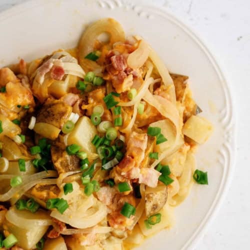 A plate of diced potatoes mixed with cooked onions, chopped bacon, melted cheese, and topped with sliced green onions.