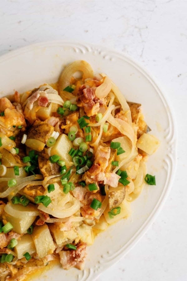 A plate of diced potatoes mixed with cooked onions, chopped bacon, melted cheese, and topped with sliced green onions.