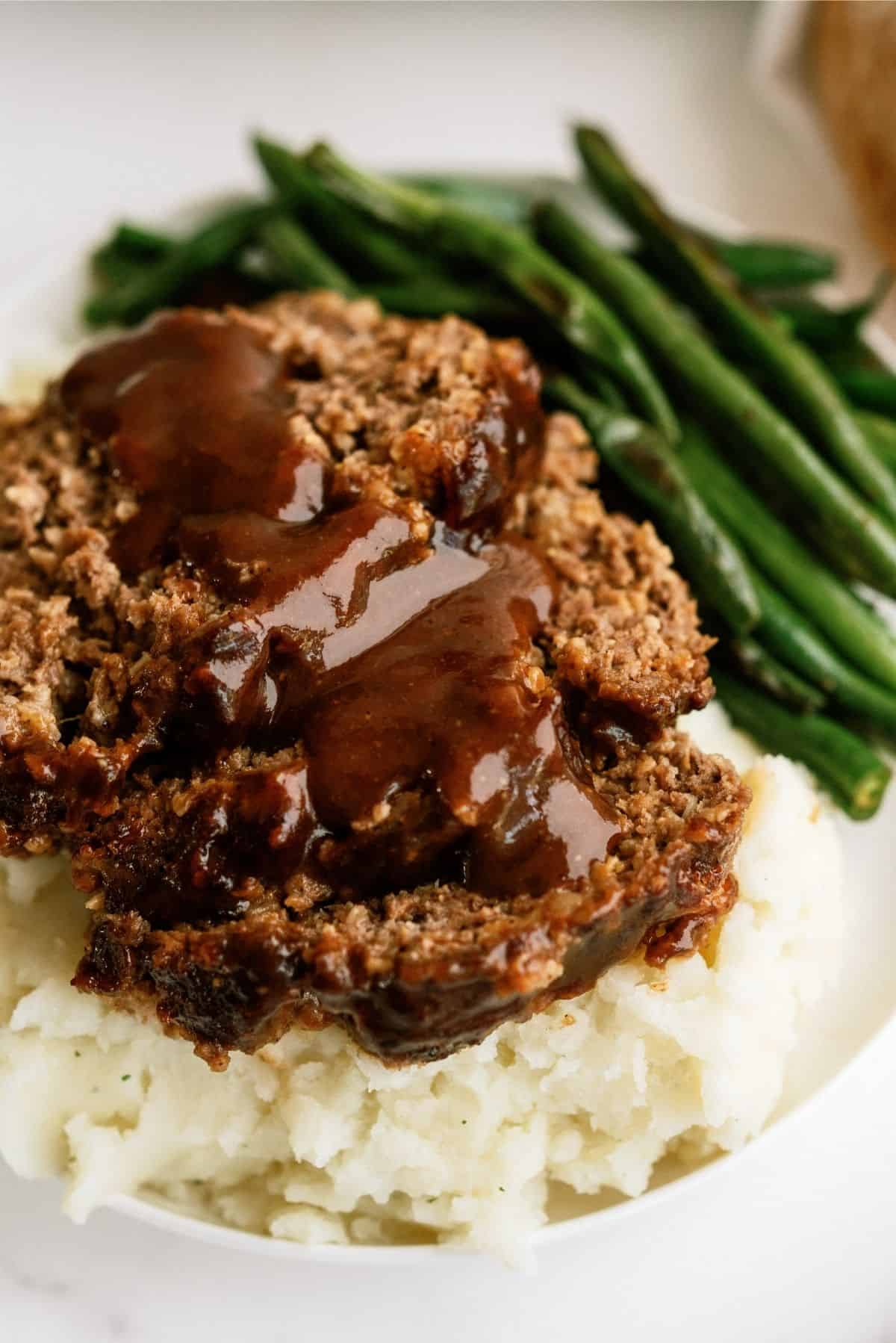 A plate of meatloaf slices topped with gravy, accompanied by mashed potatoes and green beans.