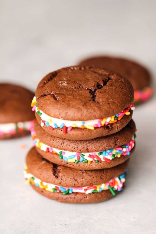 A stack of three chocolate whoopie pies with colorful sprinkles and a cream filling are arranged on a white surface.