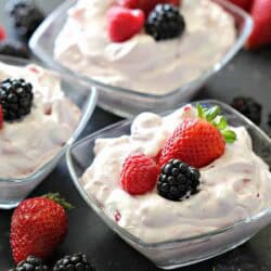 Three glass bowls filled with whipped cream and topped with strawberries, blackberries, and raspberries, surrounded by assorted fresh berries on a dark surface.