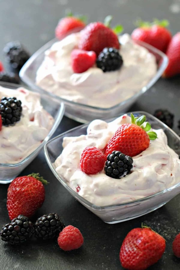 Three glass bowls filled with whipped cream and topped with strawberries, blackberries, and raspberries, surrounded by assorted fresh berries on a dark surface.
