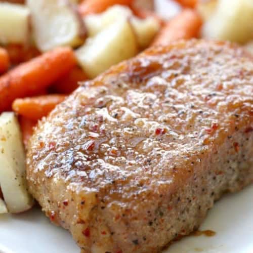 A seasoned pork chop served on a white plate with roasted potatoes and carrots in the background.