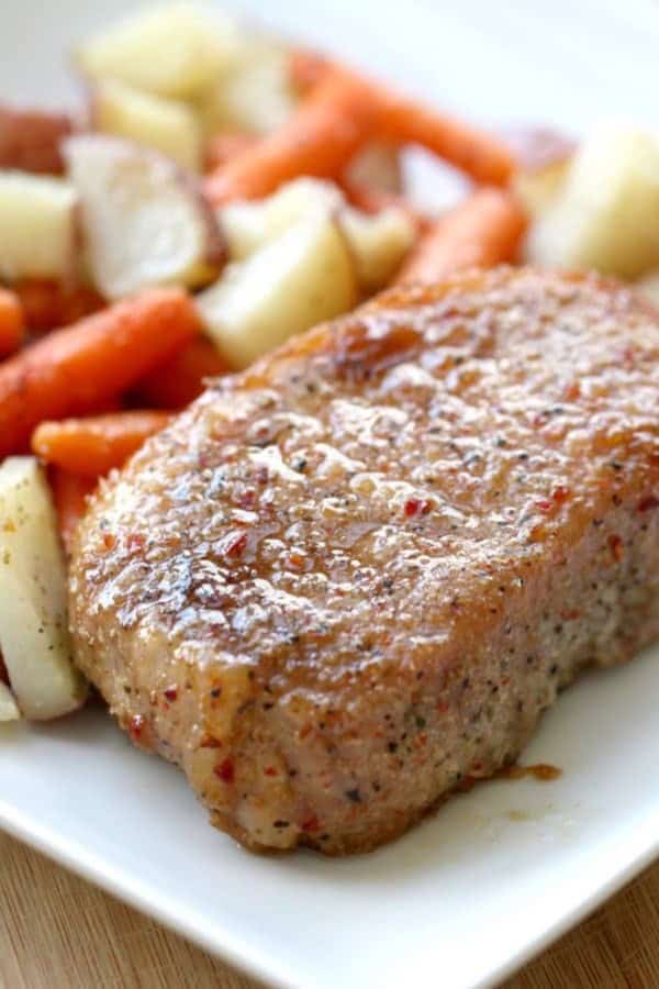 A seasoned pork chop served on a white plate with roasted potatoes and carrots in the background.