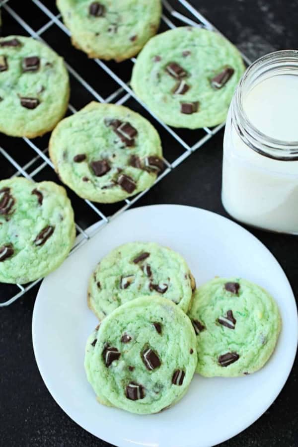 Three green cookies with chocolate chunks on a white plate, with one more green cookie on a cooling rack in the background.