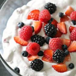 A glass bowl filled with whipped cream, topped with sliced strawberries, raspberries, blackberries, and blueberries.