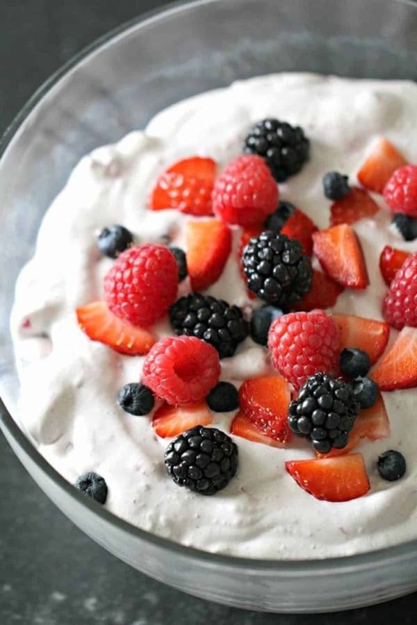 A glass bowl filled with whipped cream, topped with sliced strawberries, raspberries, blackberries, and blueberries.