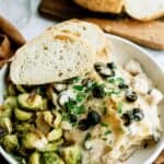 A dish with creamy pasta topped with black olives and parsley, served with roasted Brussels sprouts and two slices of bread on the side.