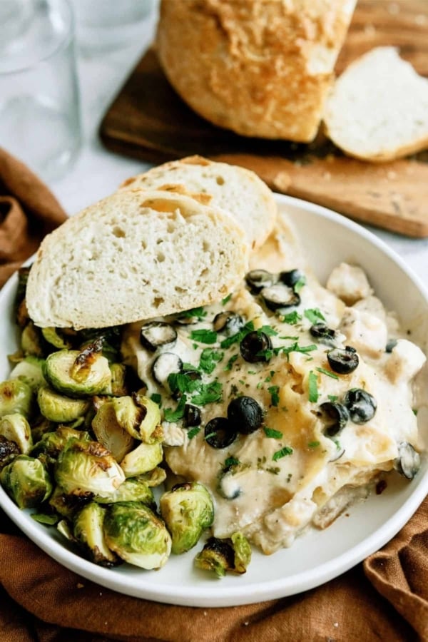 A dish with creamy pasta topped with black olives and parsley, served with roasted Brussels sprouts and two slices of bread on the side.