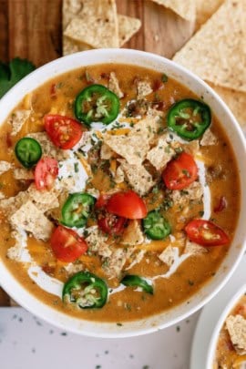 A close-up of a bowl of creamy soup garnished with sliced jalapeños, tortilla chips, and diced tomatoes.