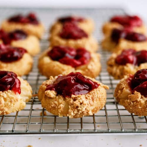 A cooling rack with twelve thumbprint cookies topped with a dark red jam filling. The cookies have a crumbly, golden-brown texture.
