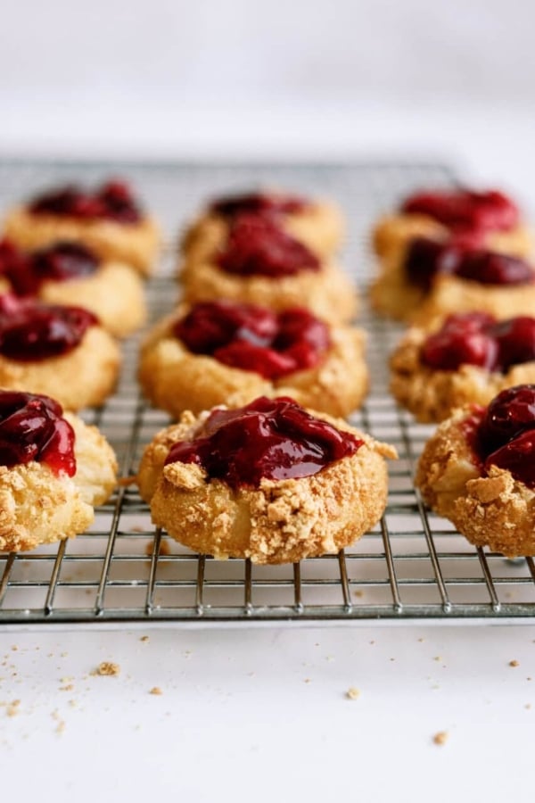 A cooling rack with twelve thumbprint cookies topped with a dark red jam filling. The cookies have a crumbly, golden-brown texture.
