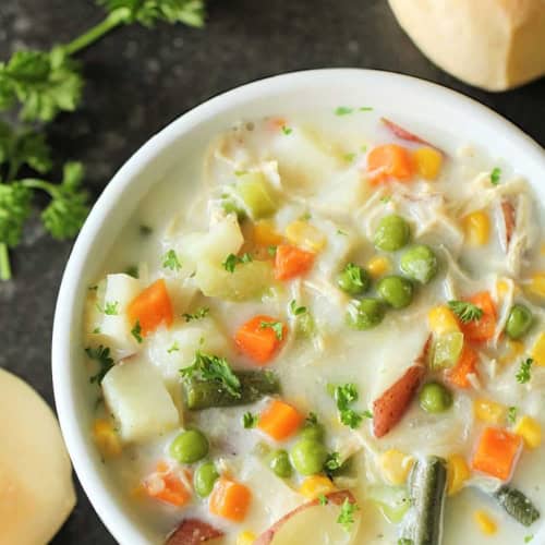 A bowl of creamy vegetable soup with potatoes, carrots, peas, and green beans, garnished with parsley, next to three bread rolls on a dark surface.