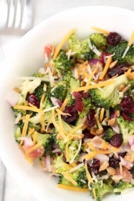 A bowl of broccoli salad topped with shredded cheese, red onions, dried cranberries, sunflower seeds, and shredded carrots.