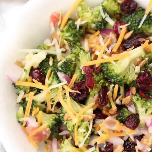 A bowl of broccoli salad topped with shredded cheese, red onions, dried cranberries, sunflower seeds, and shredded carrots.