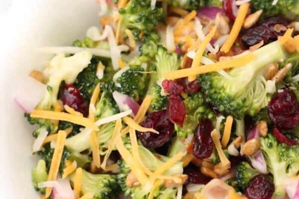 A bowl of broccoli salad topped with shredded cheese, red onions, dried cranberries, sunflower seeds, and shredded carrots.