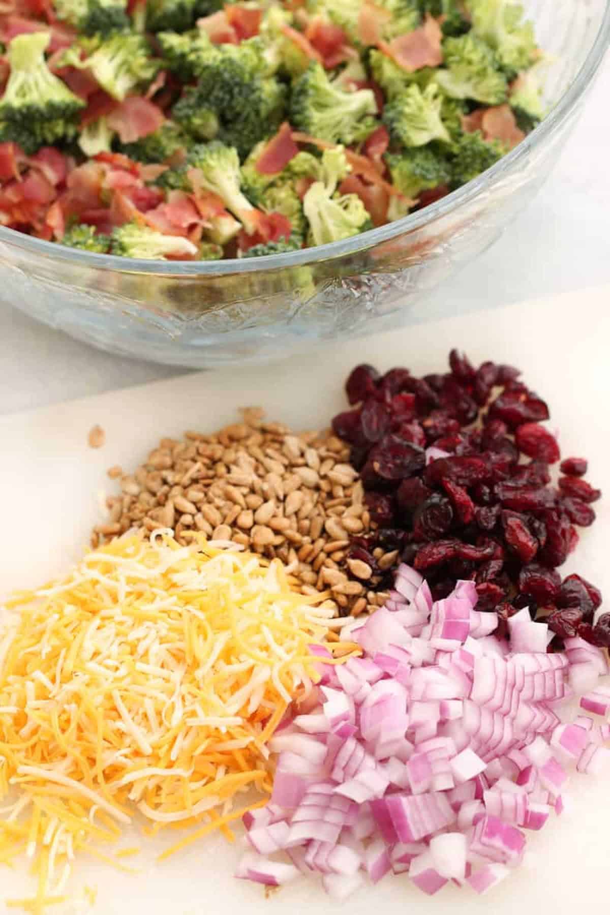 Chopped broccoli and bacon in a bowl with shredded cheese, diced red onion, dried cranberries, and sunflower seeds on a cutting board.
