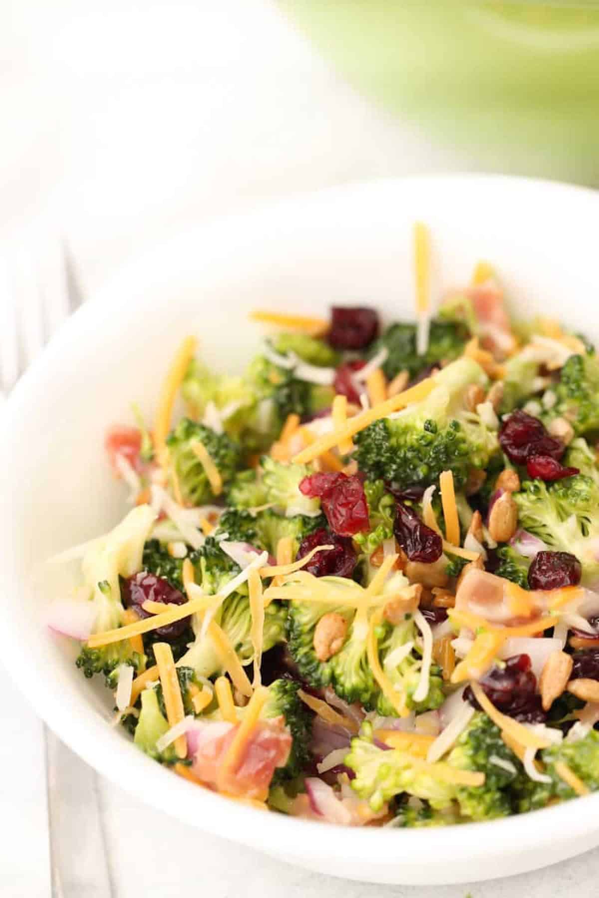 A bowl of broccoli salad with shredded cheese, dried cranberries, sunflower seeds, red onion, and diced ham.