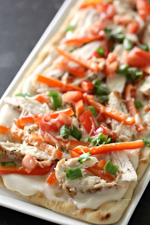 Close-up of a flatbread topped with sliced grilled chicken, diced tomatoes, red bell peppers, green onions, and melted cheese on a rectangular white plate.