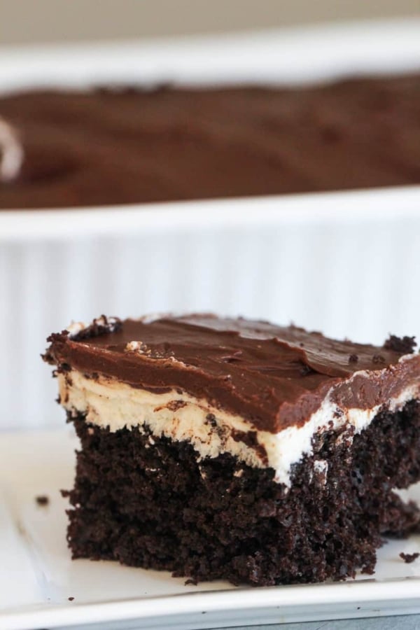 Close-up of a piece of layered chocolate cake with a creamy middle and chocolate icing on a white plate. The background shows a baking dish with more cake.