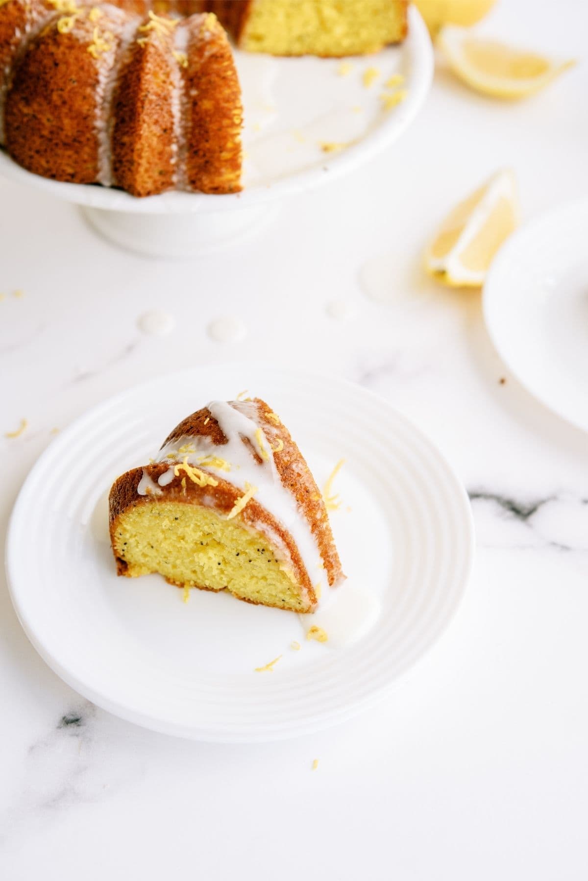 A slice of Lemon Poppy Seed Bundt Cake on a plate with the remaining Bundt cake in the background on a cake stand.