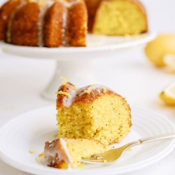 A slice of Lemon Poppy Seed Bundt Cake on a white plate with the remaining bundt cake on a cake stand in the background.