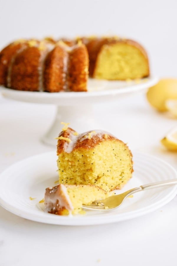 A slice of Lemon Poppy Seed Bundt Cake on a white plate with the remaining bundt cake on a cake stand in the background.