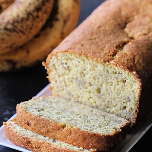 3 Ingredient Banana Bread on a platter with some of the loaf sliced, ripe bananas in the background.