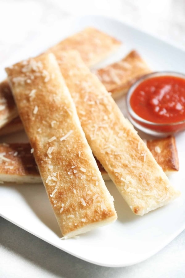 A square white plate holds several breadsticks, lightly dusted with cheese, served alongside a small dish of marinara sauce.
