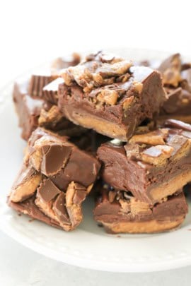A white plate contains a stack of chocolate and peanut butter fudge squares, each topped with pieces of candy.