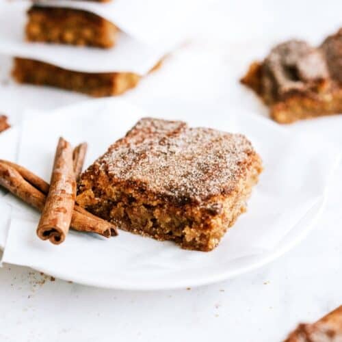 One Snickerdoodle Blondie on a plate with a cinnamon stick. Other Snickerdoodle Blondies on plates in the background.