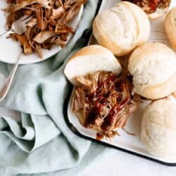 A plate of shredded meat next to a tray of sandwich rolls filled with meat and topped with barbecue sauce, set on a light surface with a green cloth.