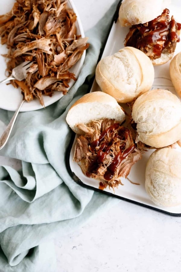 A plate of shredded meat next to a tray of sandwich rolls filled with meat and topped with barbecue sauce, set on a light surface with a green cloth.
